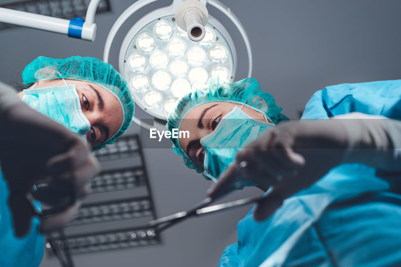 From below female surgeons in medical uniform using professional tools while standing under bright light in operating theater