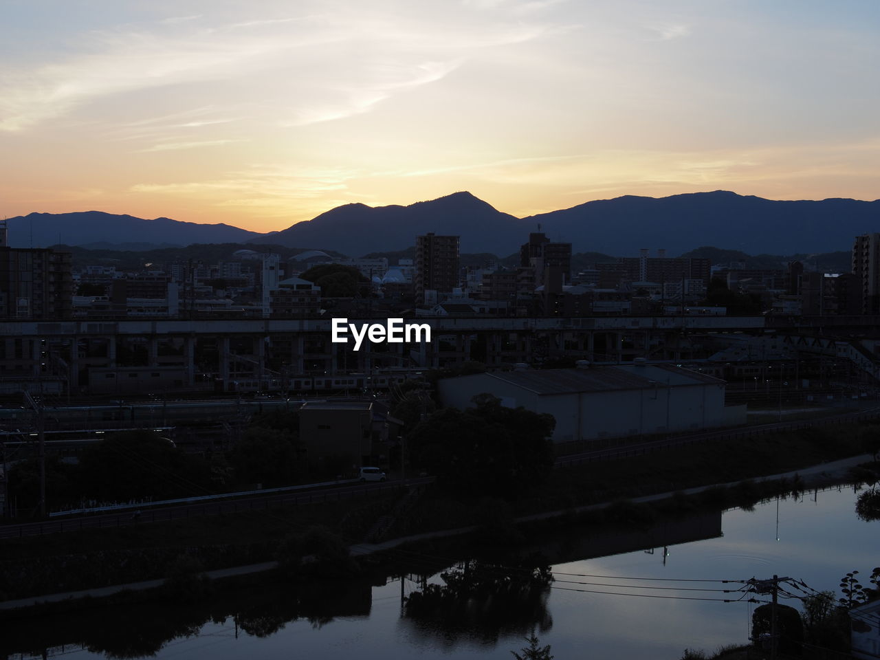 REFLECTION OF SILHOUETTE BUILDINGS AND RIVER AGAINST SKY DURING SUNSET