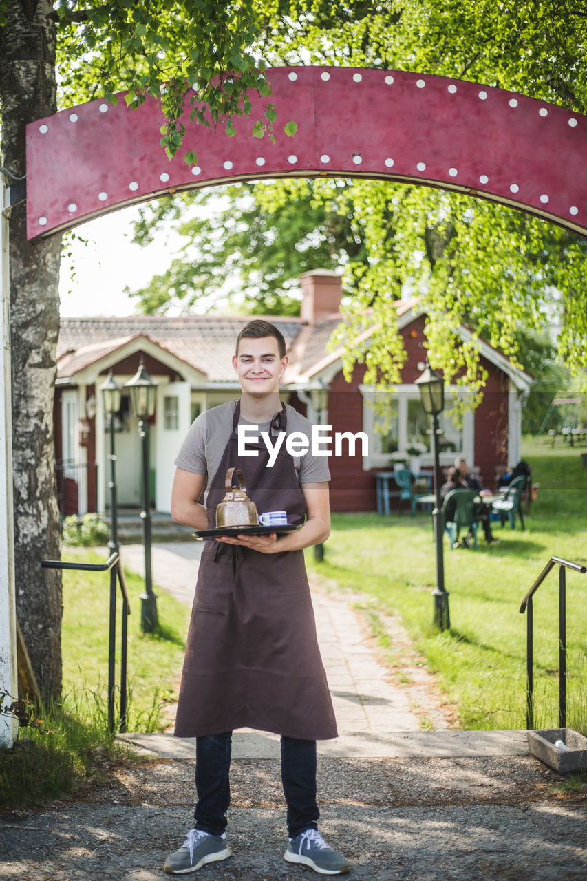 Full length portrait of smiling young waiter holding serving tray while standing at outdoor cafe entrance