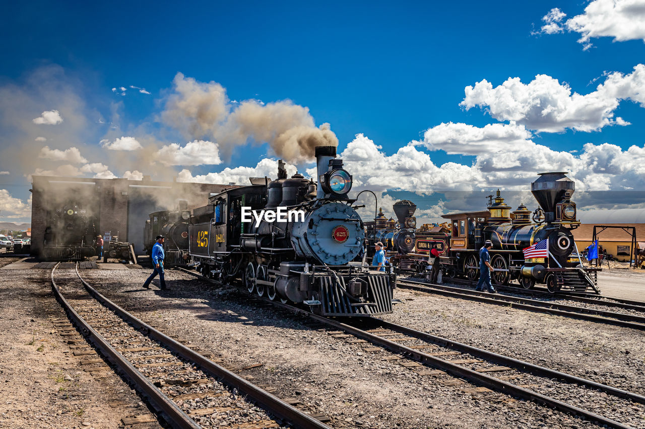 rail transportation, railroad track, track, sky, train, transportation, cloud, mode of transportation, transport, railway, locomotive, vehicle, smoke, nature, public transportation, steam train, day, travel, sunlight, steam, outdoors, land vehicle, business, group of people, steam engine, railroad station