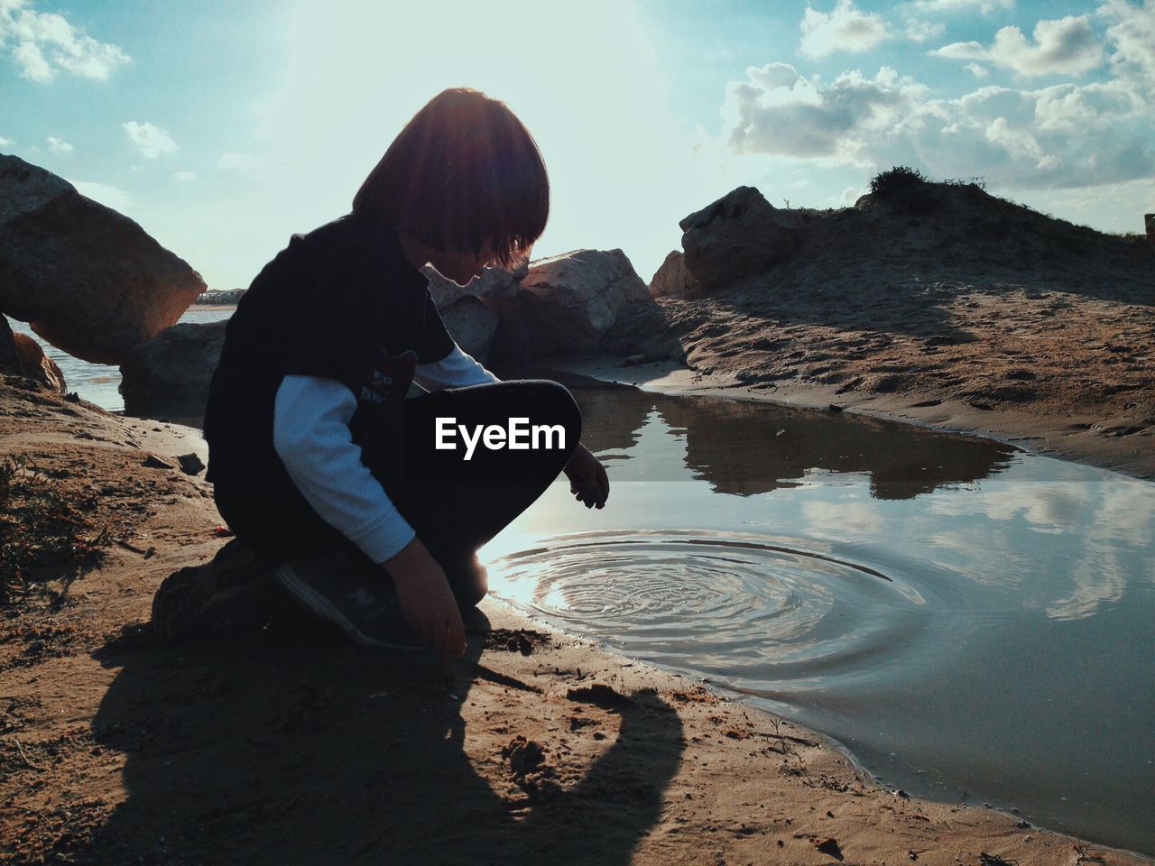 Side view of boy crouching by puddle against sky during sunny day