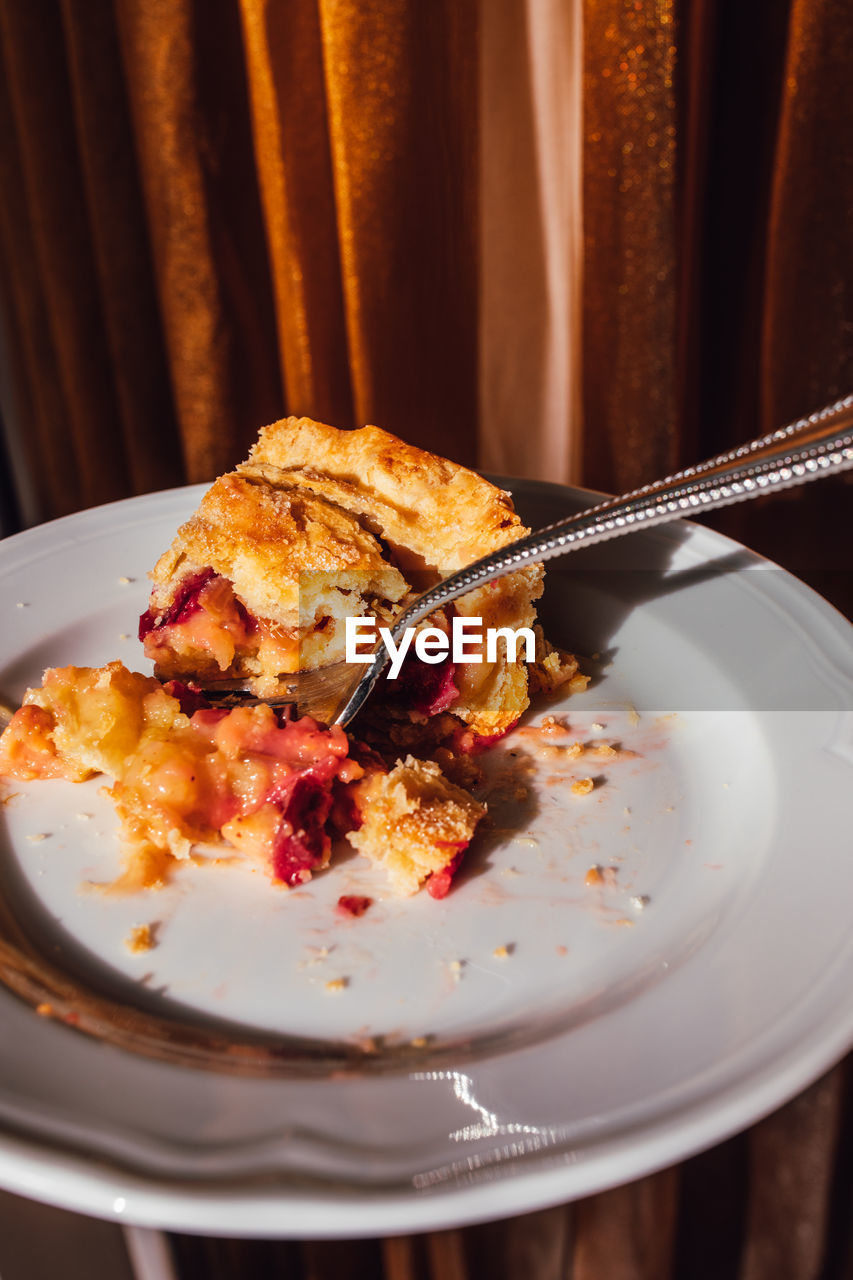 Half eaten slice of rhubarb custard pie on white plate with fork with golden curtain background