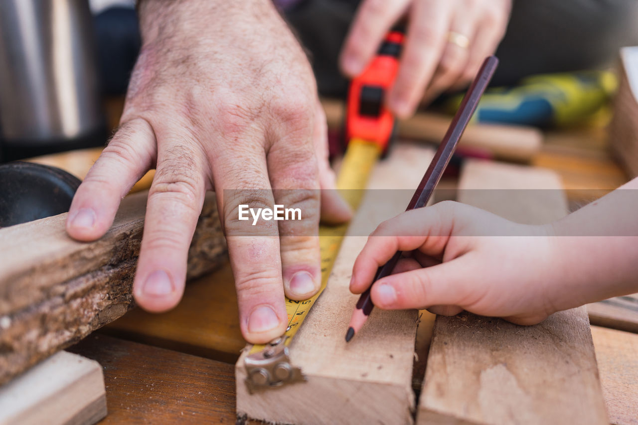 Crop unrecognizable dad measuring wooden piece with tape against child with pencil sitting on boardwalk