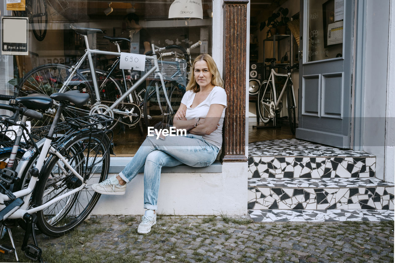 Portrait of confident female owner with arms crossed sitting against bicycle shop