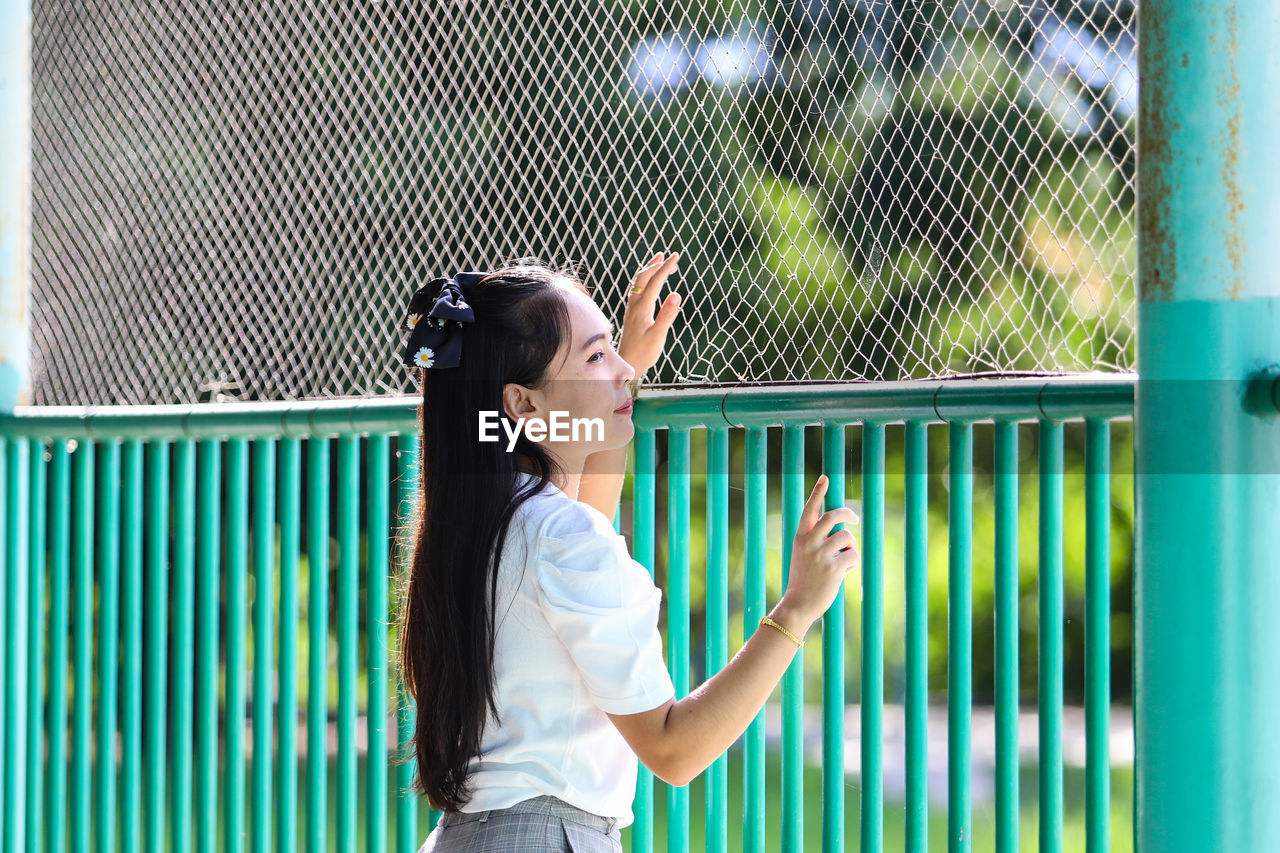 Side view of young woman standing against fence