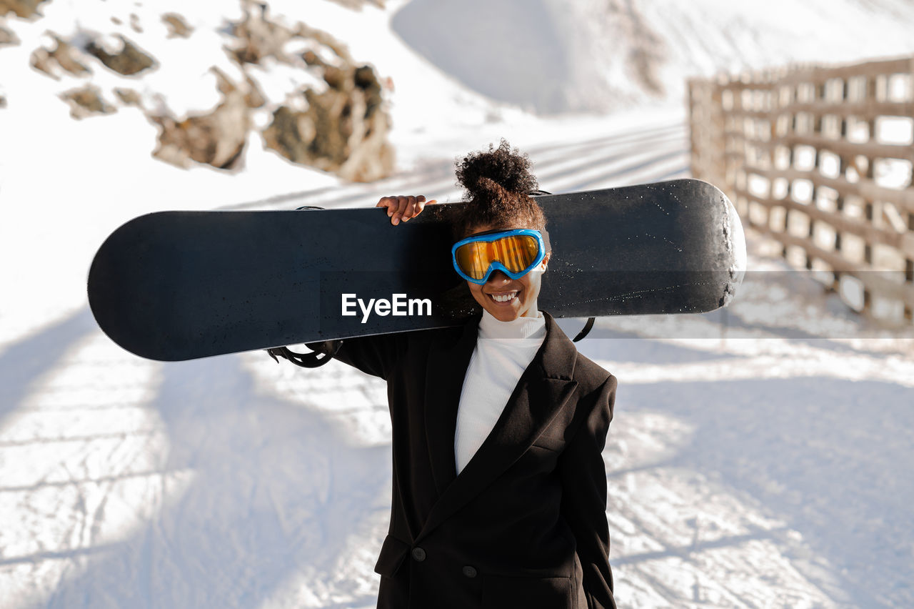 African american woman with goggles and a snowboard on a snowy mountain during winter