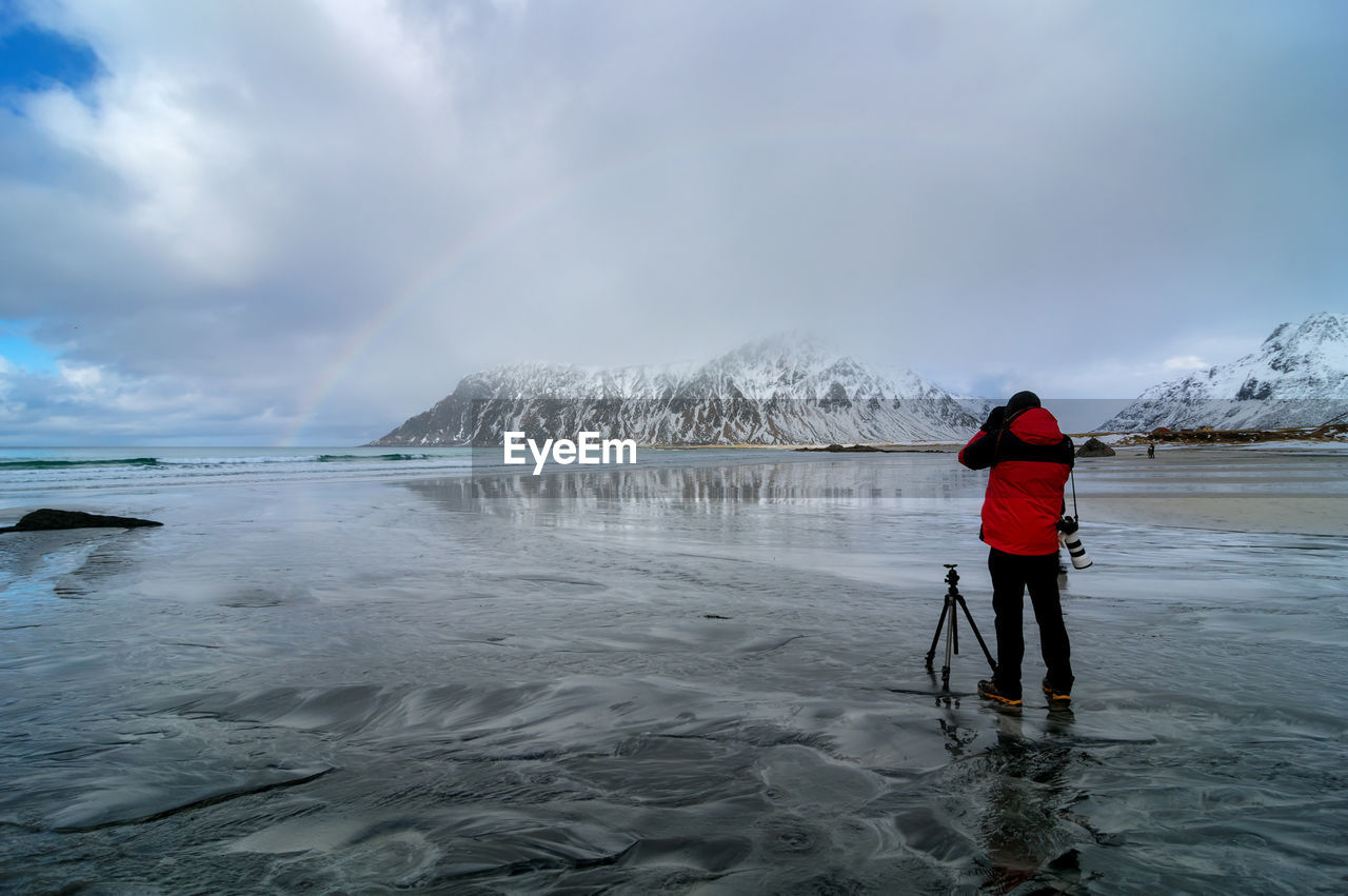 Full length of man photographing rainbow while standing at beach against sky