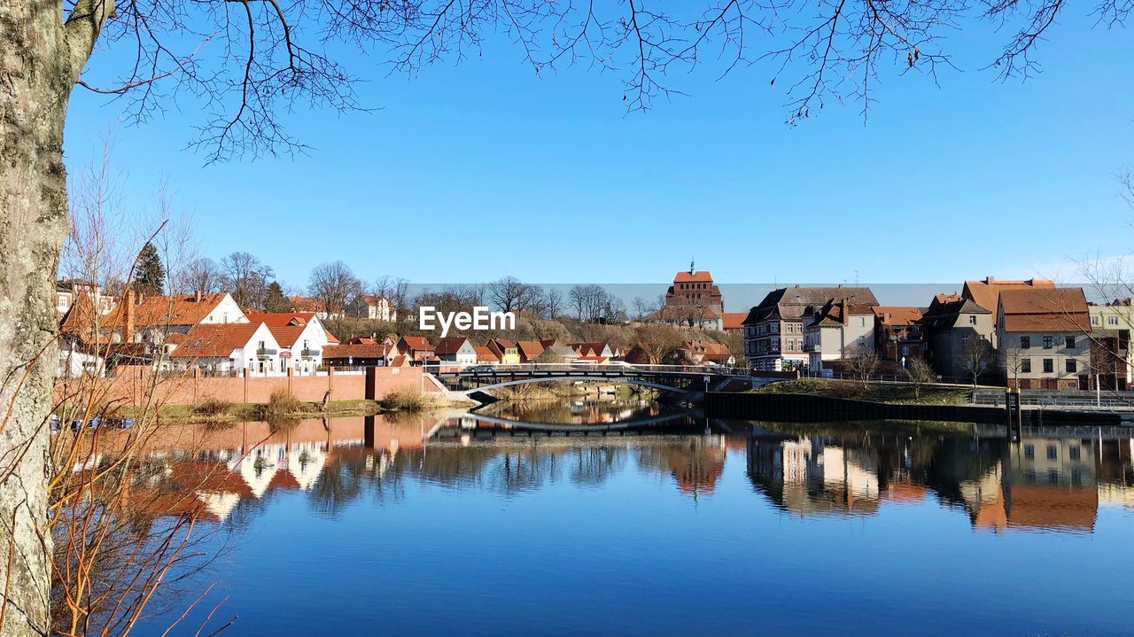 Reflection of buildings and trees in river against clear blue sky