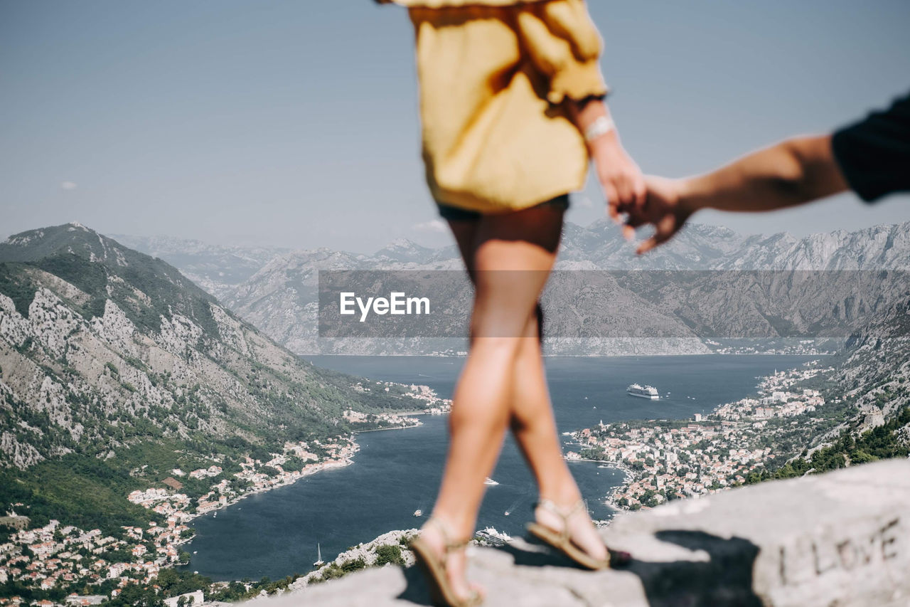 Low section of woman holding man hand while walking on retaining wall against mountain