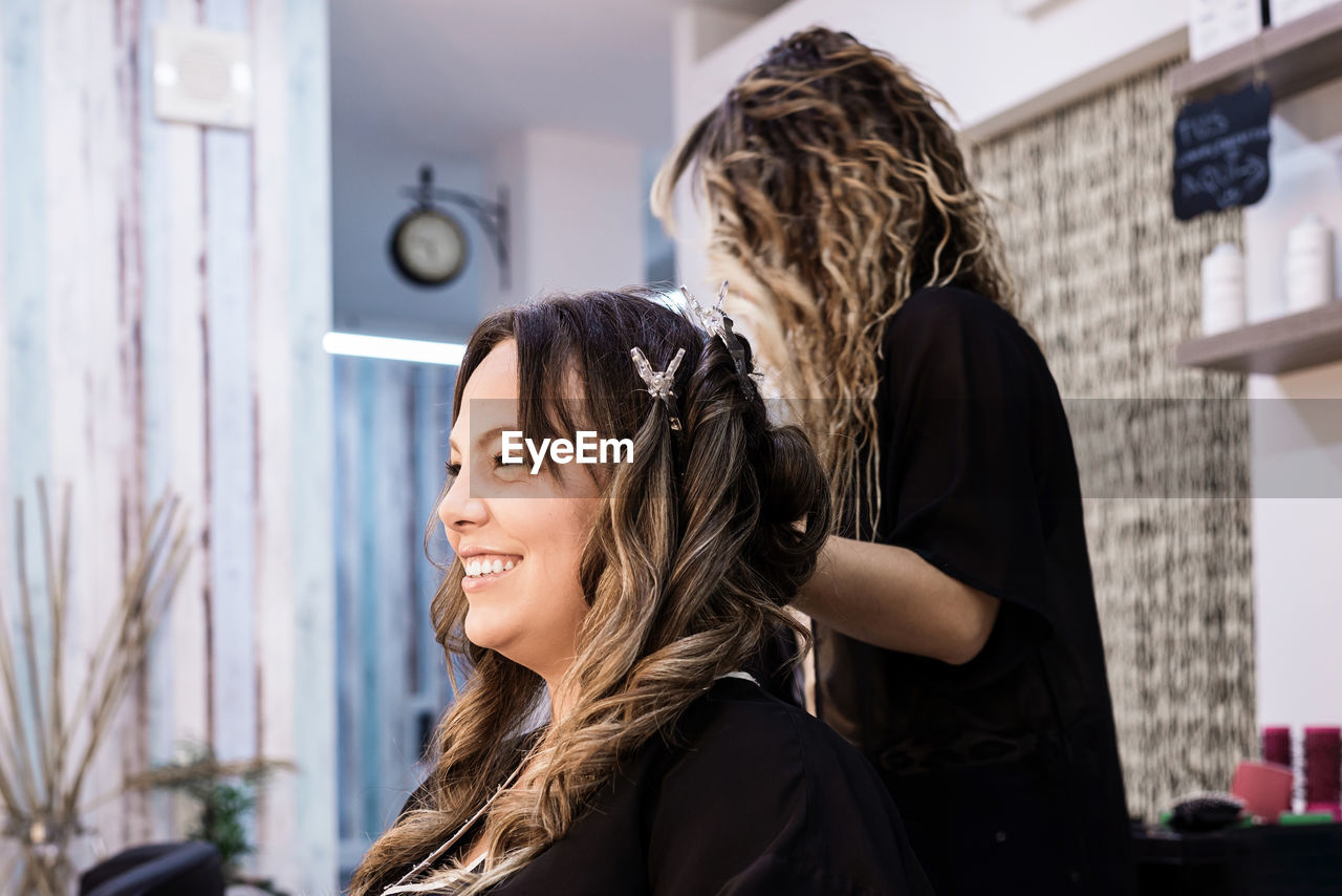 Hairdresser combing client with long curly hair against wall mirror at hair salon