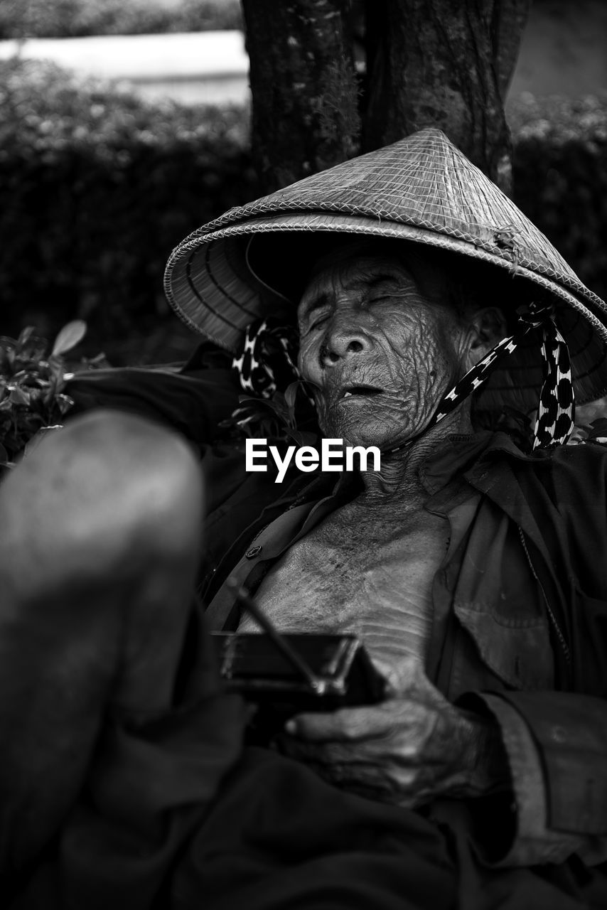 Senior man wearing asian style conical hat while napping outdoors with radio