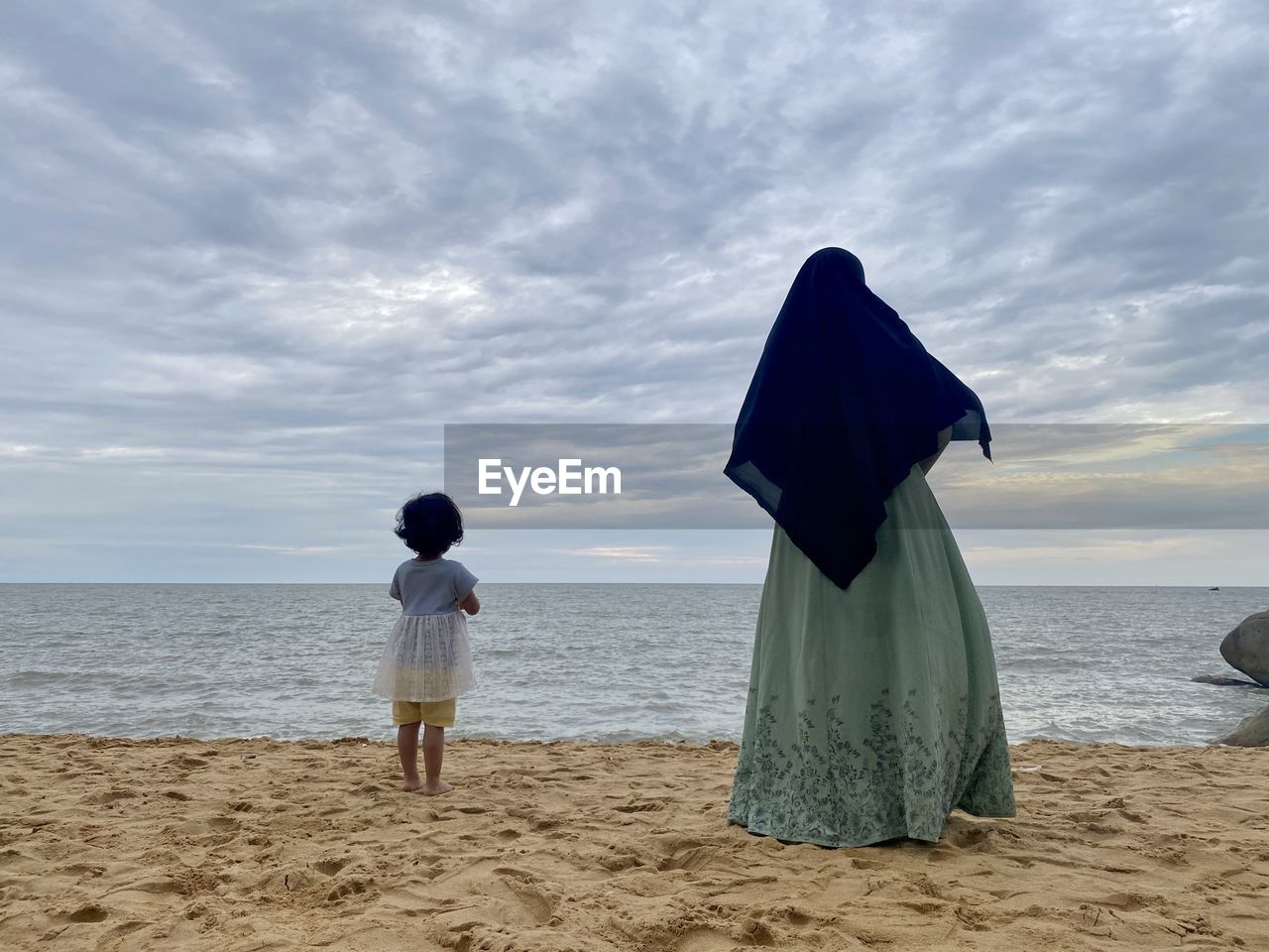 REAR VIEW OF WOMAN STANDING ON BEACH AGAINST SKY