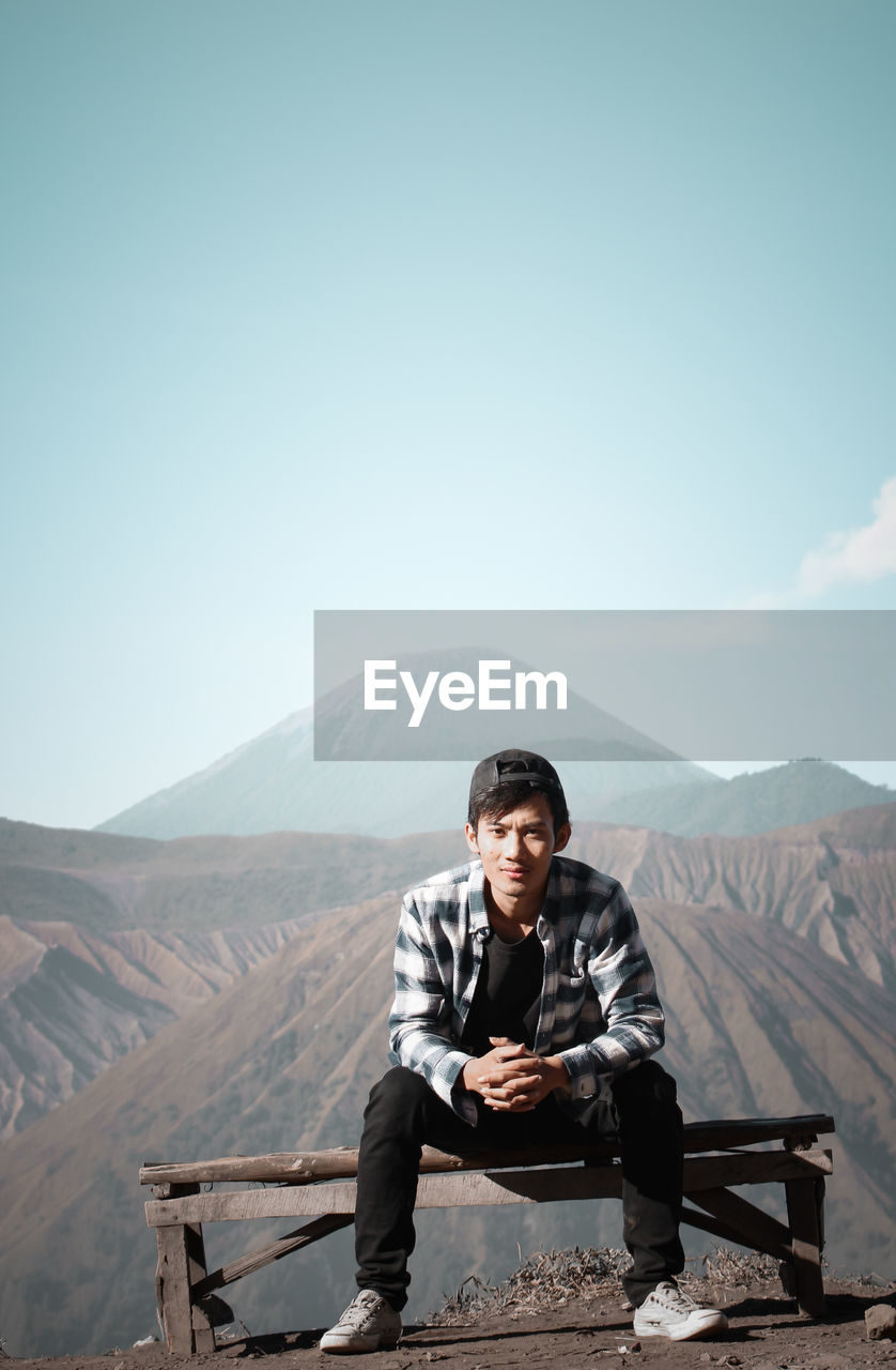 Portrait of young man sitting on bench against mountains during sunny day