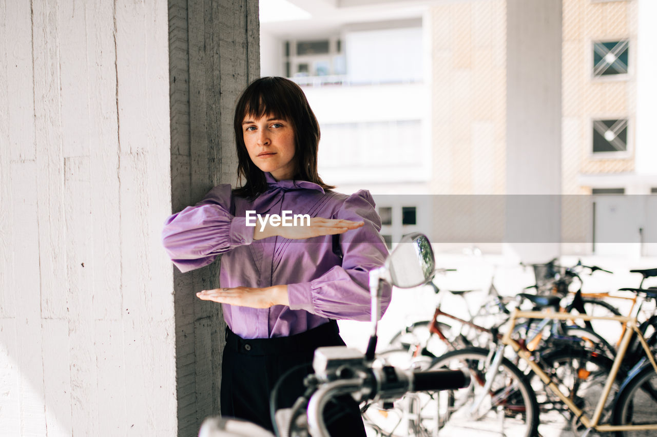 Portrait of young woman gesturing equal sign while standing by bicycle and wall