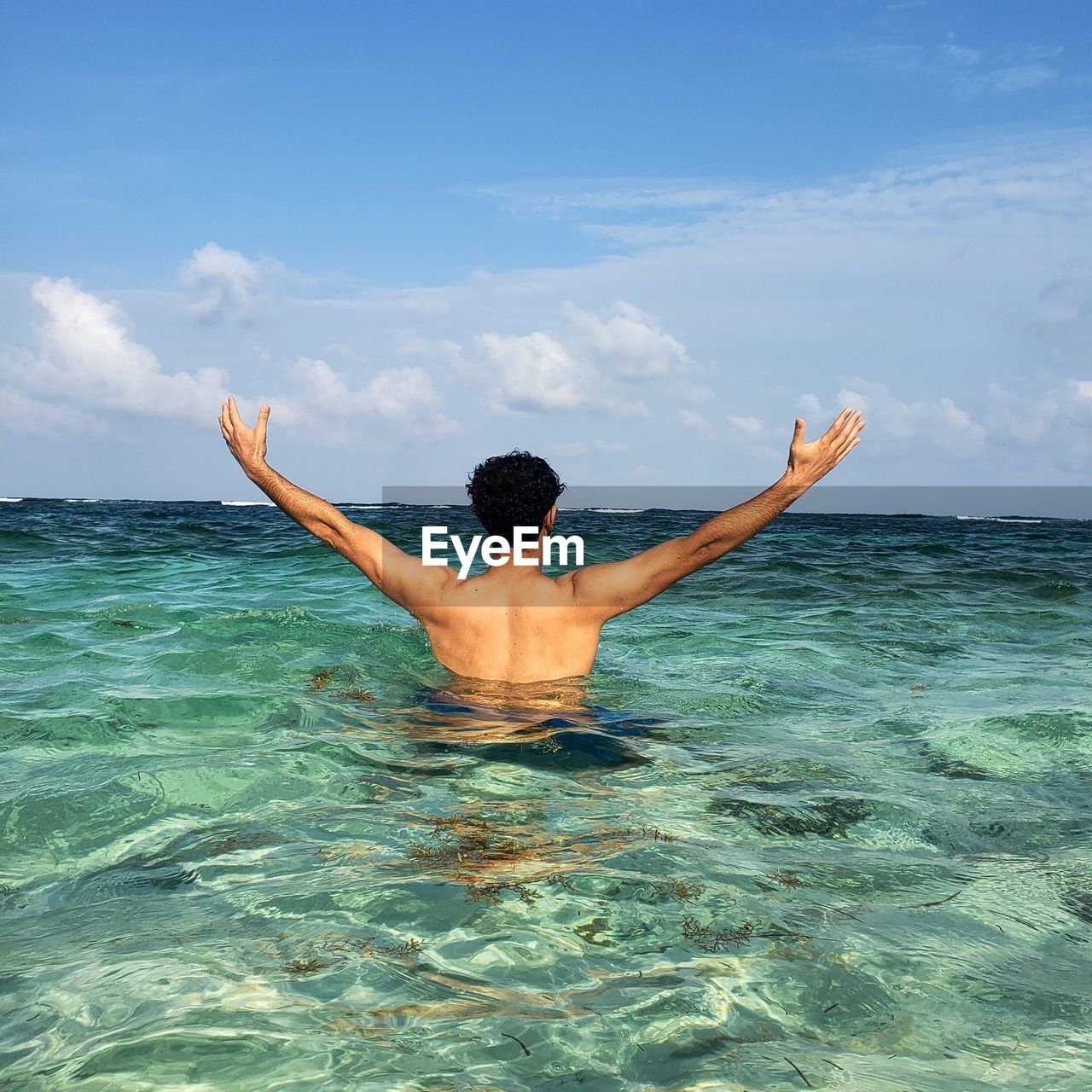 Rear view of shirtless man with arms outstretched standing in sea against sky