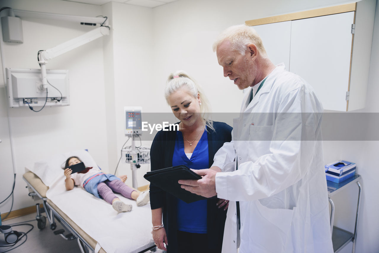 Mature doctor showing digital tablet to woman standing by bed at hospital