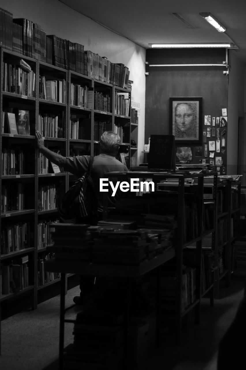 REAR VIEW OF MAN SITTING ON BOOK IN LIBRARY