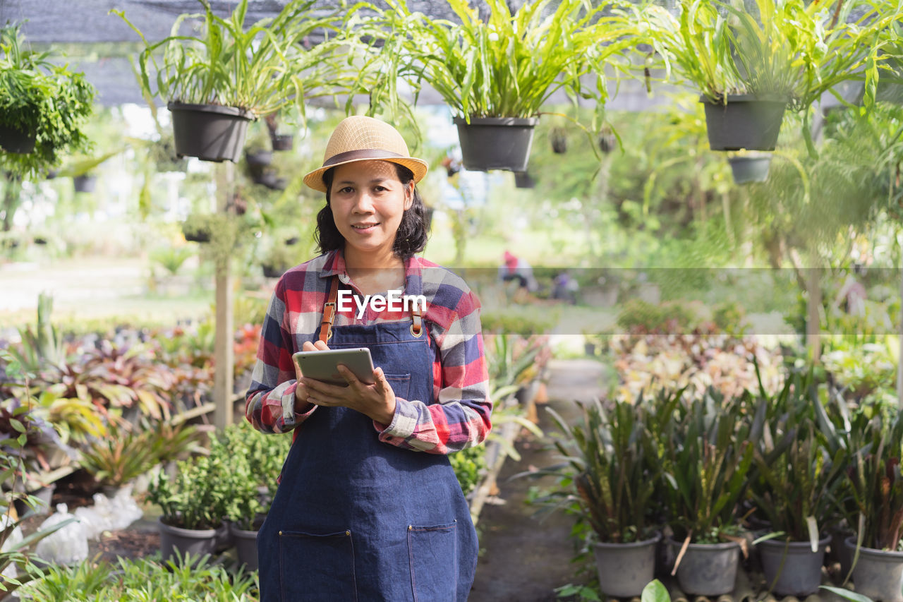 Portrait of smiling woman holding digital tablet while standing against plants