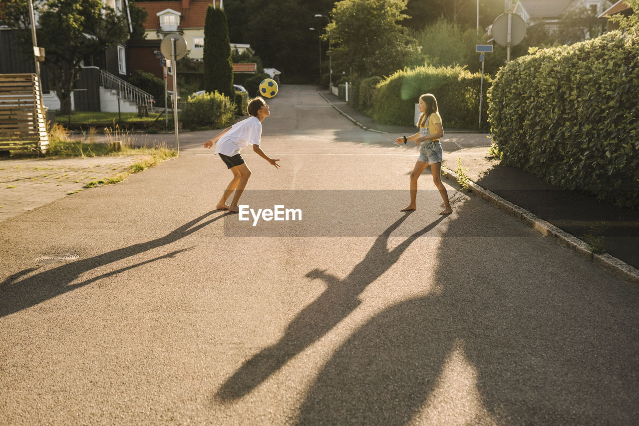 Siblings playing with soccer ball on road during sunny day