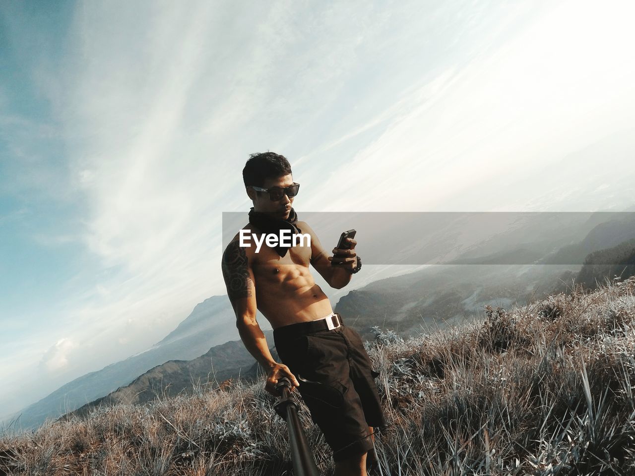 Shirtless mid adult man using mobile phone while standing on mountain against sky