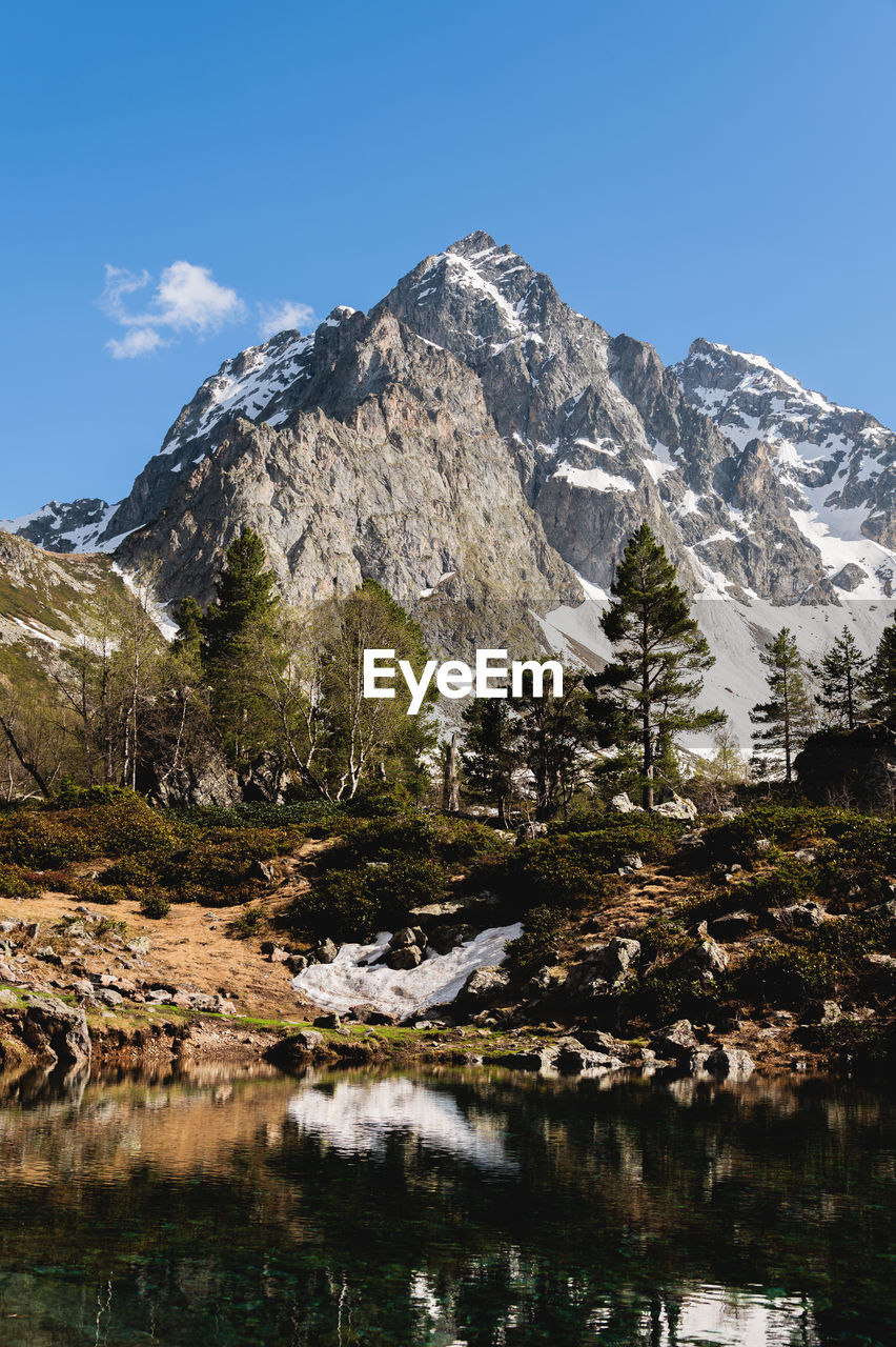 Rocky snow-capped mountains reflected in a pond or lake, forest and rocks around. clear sunny