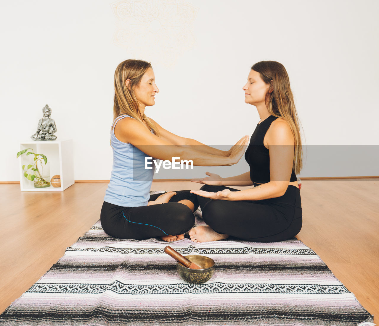 women, adult, physical fitness, sitting, two people, lifestyles, indoors, female, wellbeing, yoga, person, sports, young adult, human leg, exercising, togetherness, limb, smiling, sports clothing, full length, happiness, arm, relaxation, clothing, side view, home interior, stretching, blond hair, friendship, emotion, leisure activity, bonding, posture