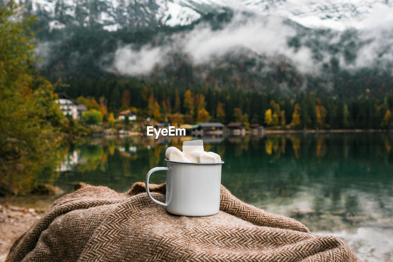 Picnic in bavarian mountains, germany. hand with mug of cacao and marshmallow on the lake background
