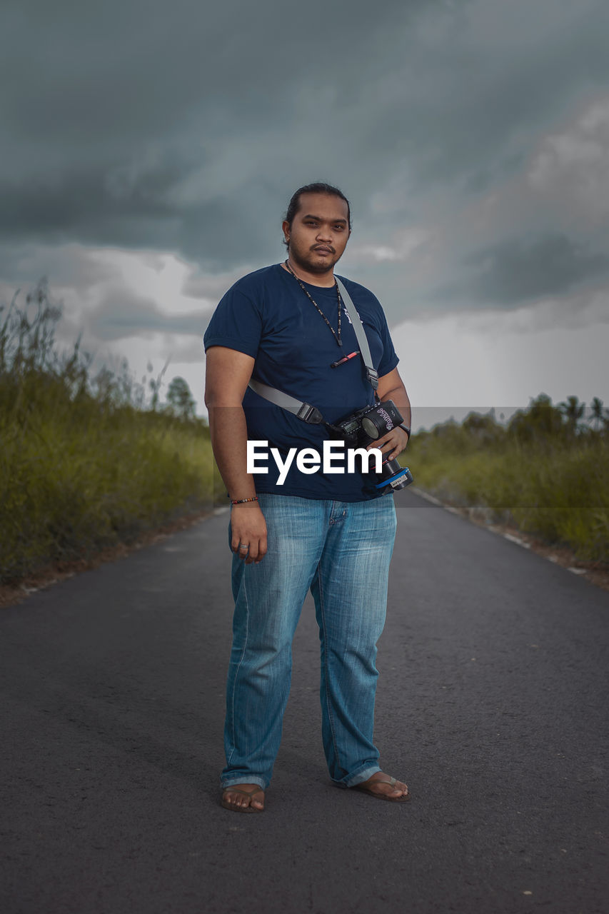 Full length portrait of young man standing on road against sky