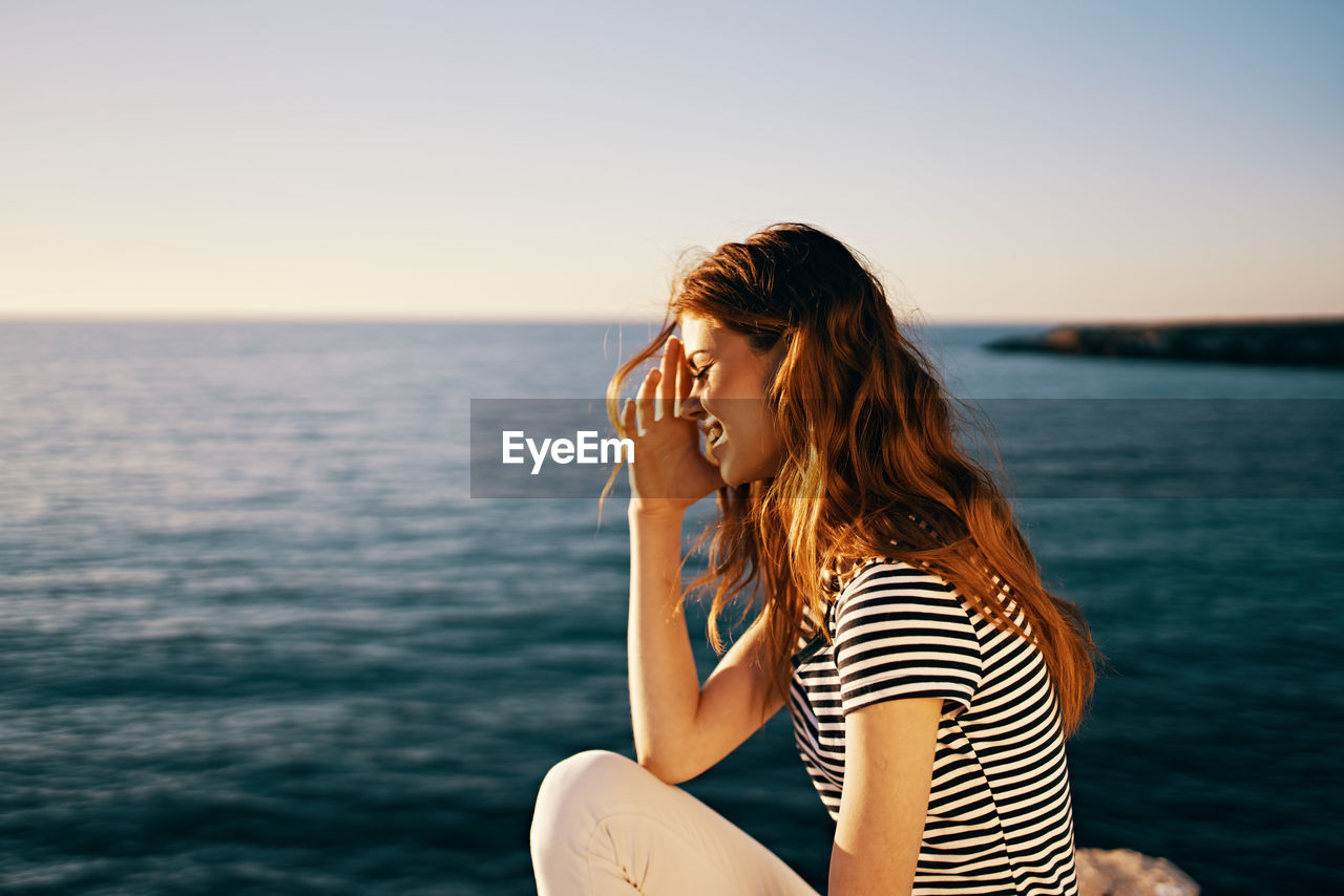 Young woman sitting by sea against sky during sunset