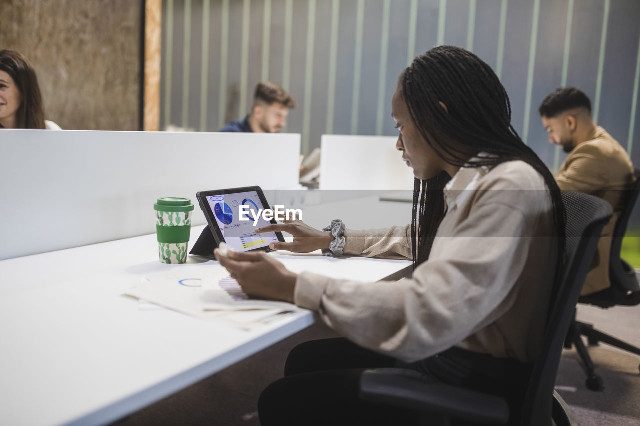 Side view of concentrated african american female employee using tablet while sitting at table and working in coworking space with multiracial colleagues
