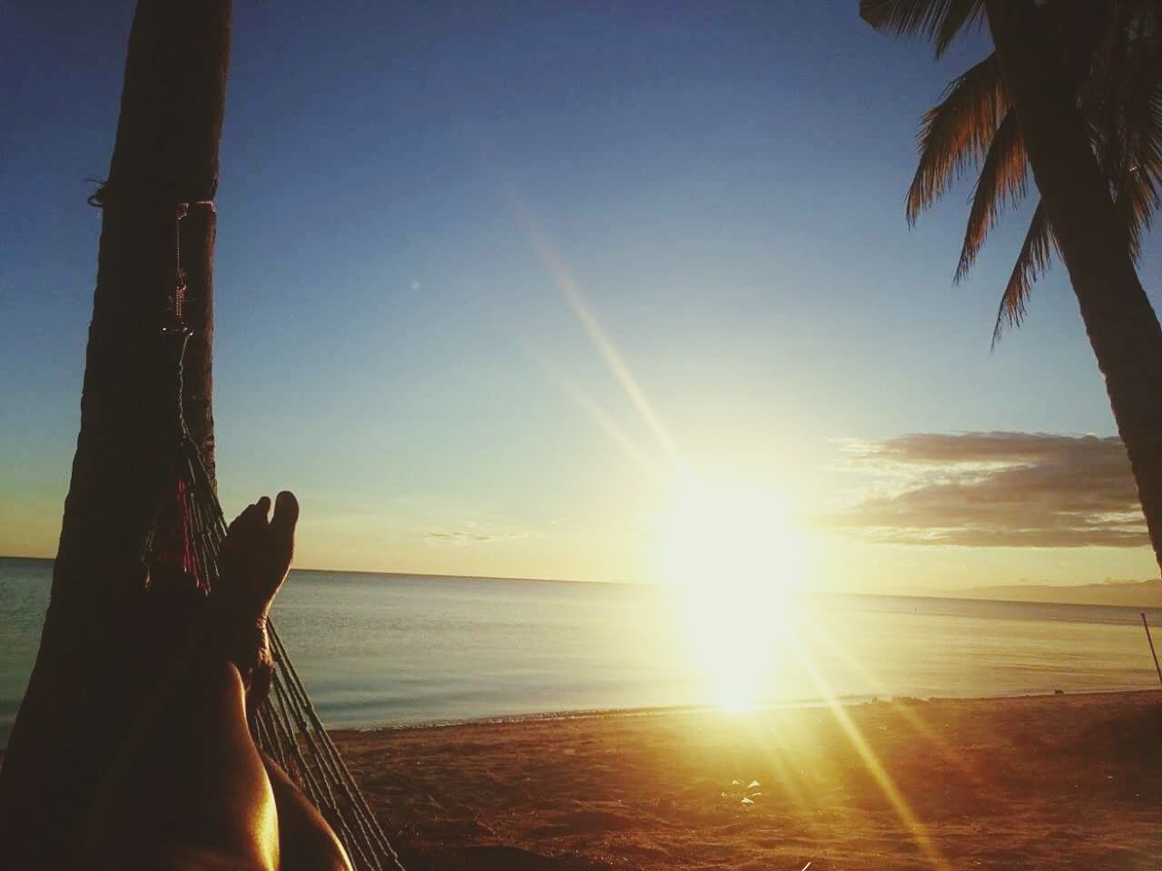 Low section of woman relaxing on hammock at beach against sky during sunset