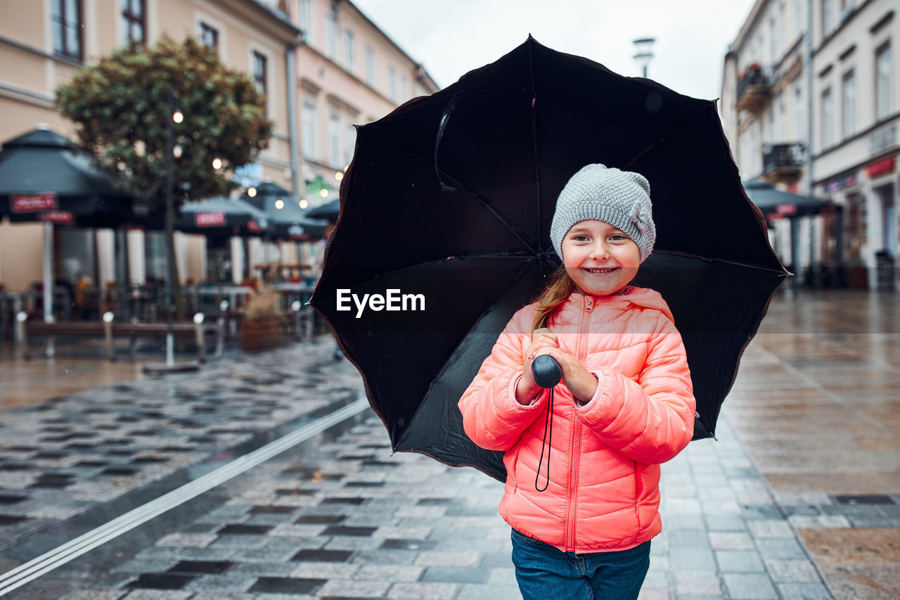 Little smiling happy girl holding big umbrella walking in a downtown on rainy gloomy autumn day