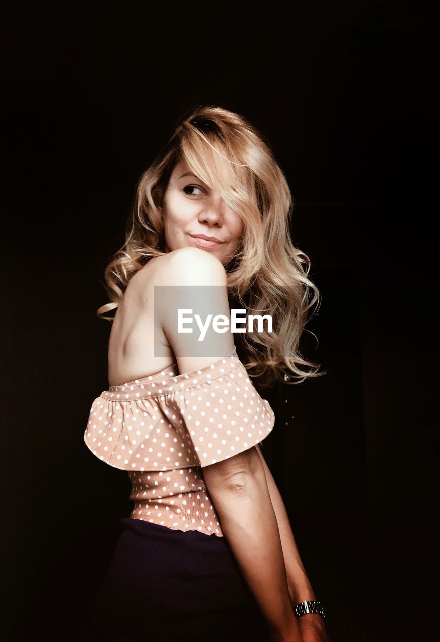 Side view of young woman with blond hair standing against black background