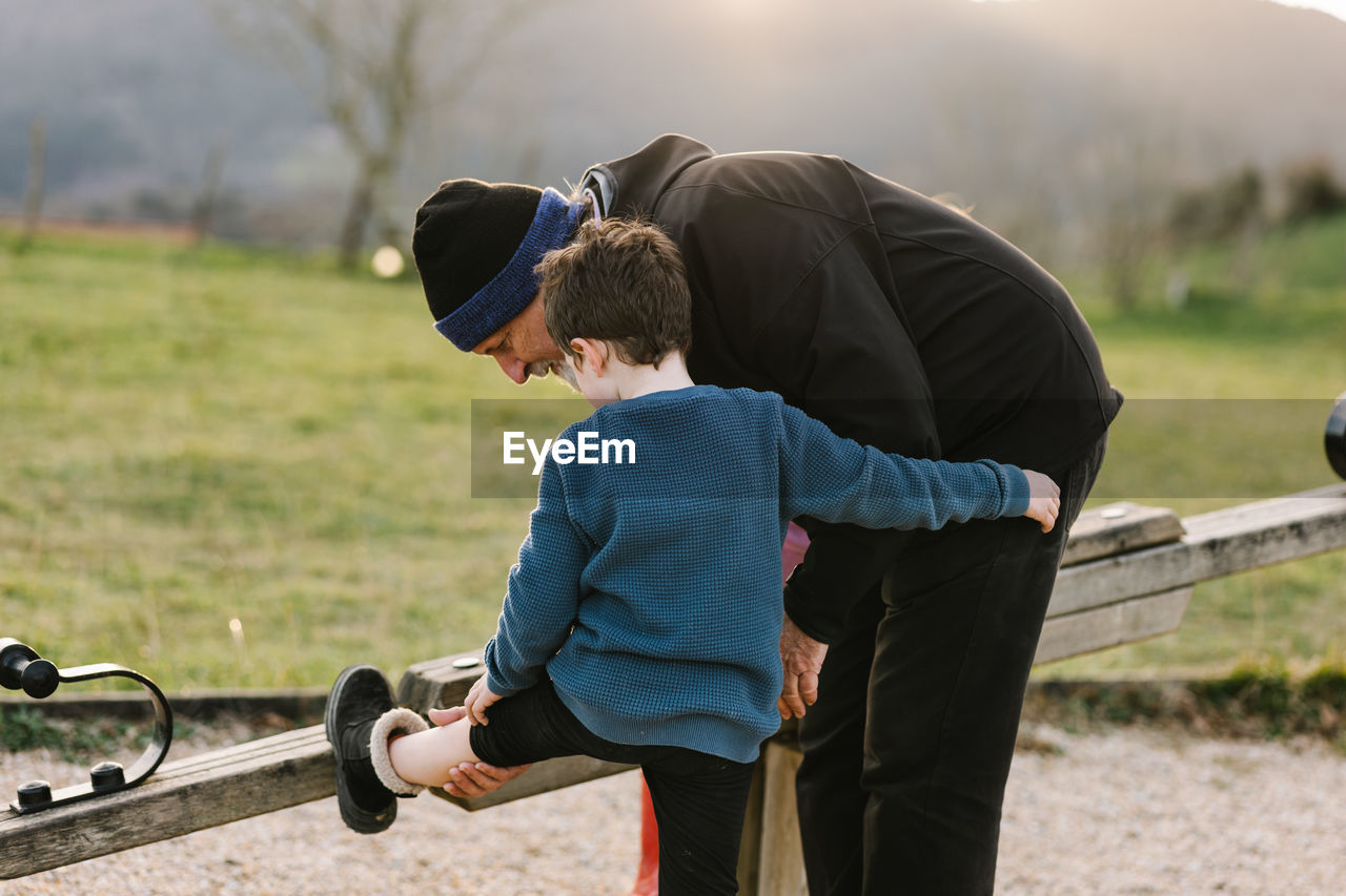 Caring grandfather looking at leg of grandson near wooden seesaw while playing on playground in countryside against mountain ridge on summer day