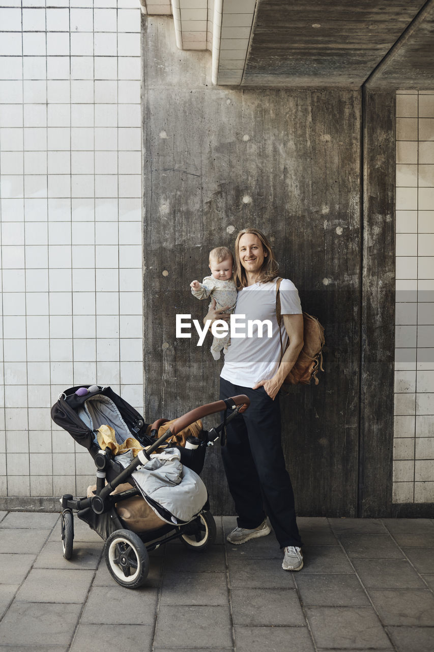 Smiling man with hand in pocket carrying daughter while standing by stroller in front of wall