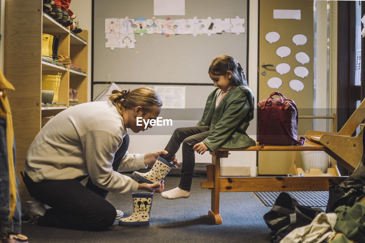 Male teacher helping girl to put on rubber boot while sitting in day care center