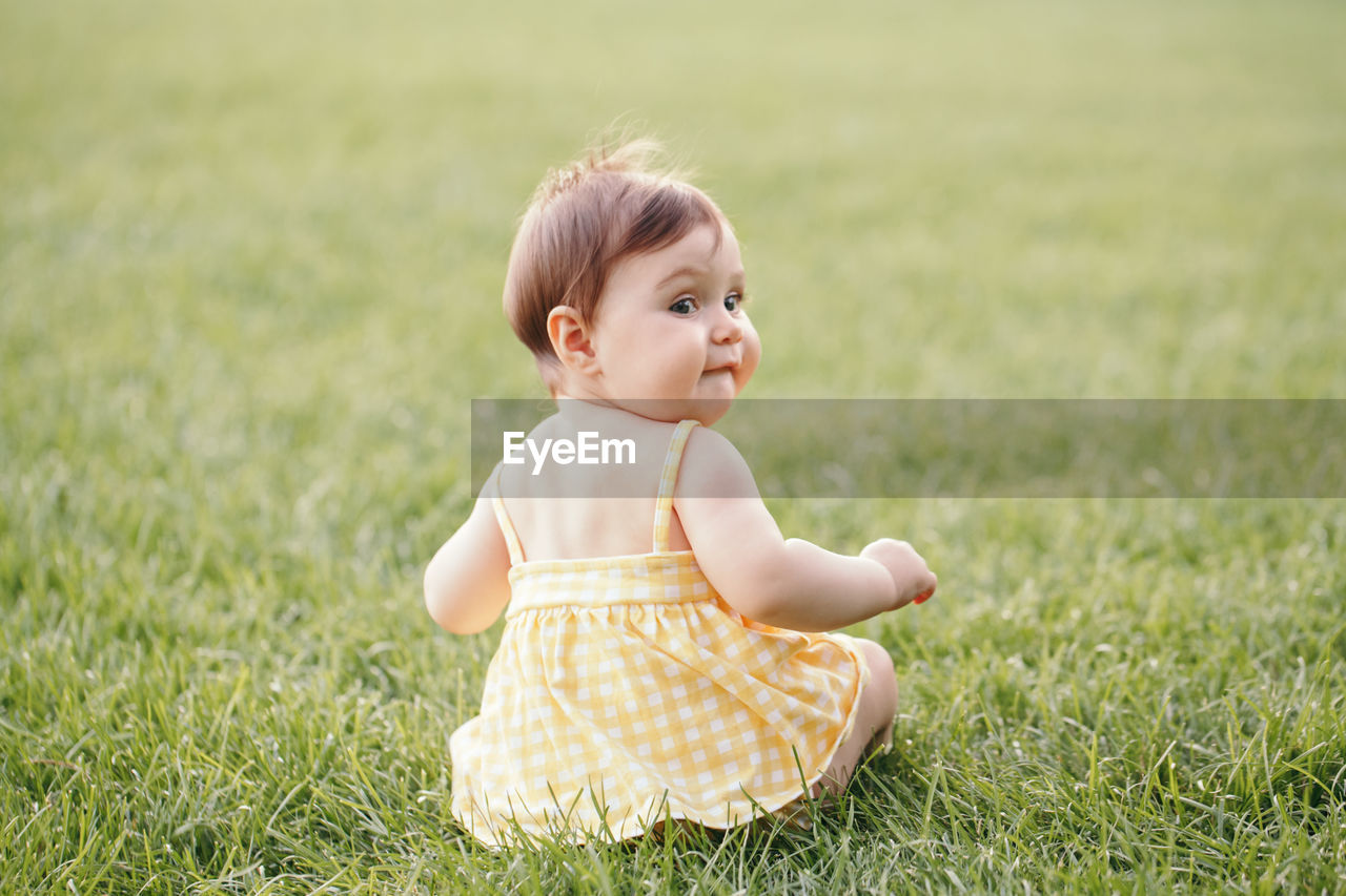 Funny baby kid. cute baby girl in yellow dress sitting on grass in park outdoor and looking back