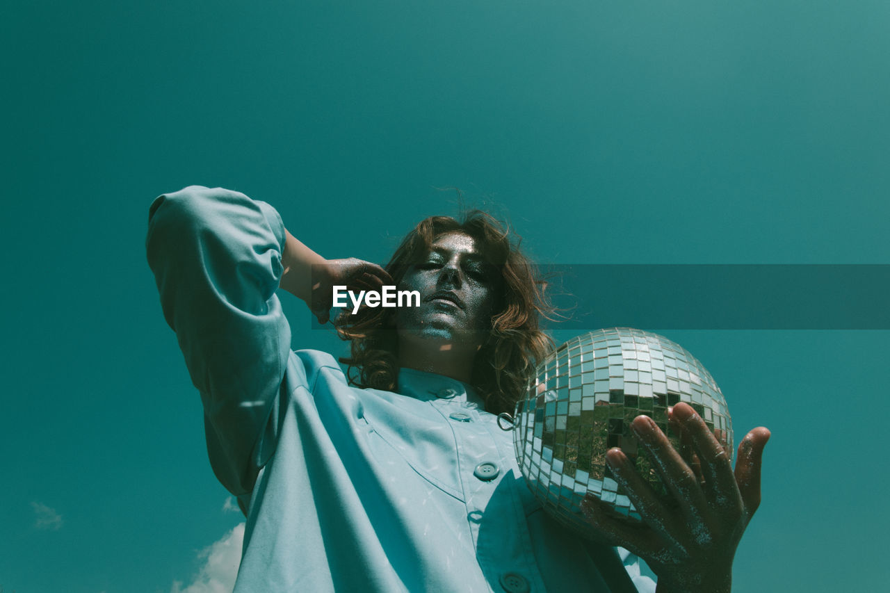 Low angle portrait of woman with glitter on face holding disco ball against clear sky