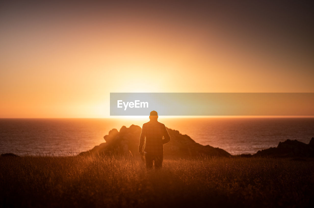 Rear view of man standing on beach against sky during sunset