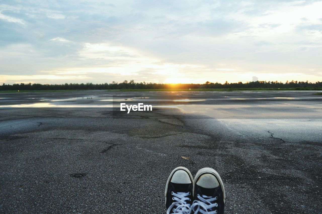 Low section of man sitting on road against cloudy sky during sunset