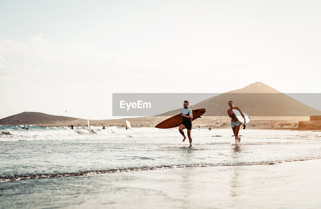 Man and woman running while holding surfboards at beach against sky