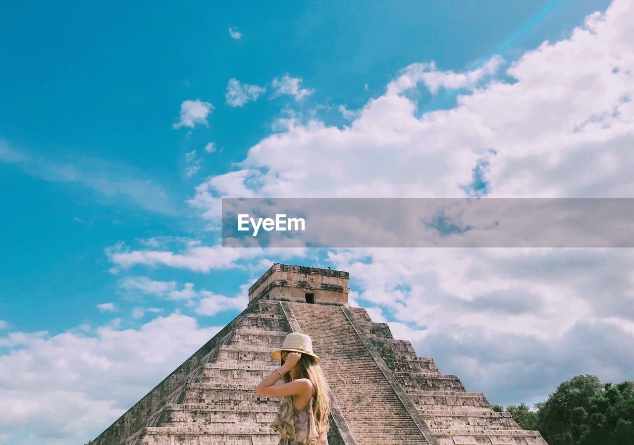 Low angle view of young woman standing by pyramid against cloudy sky