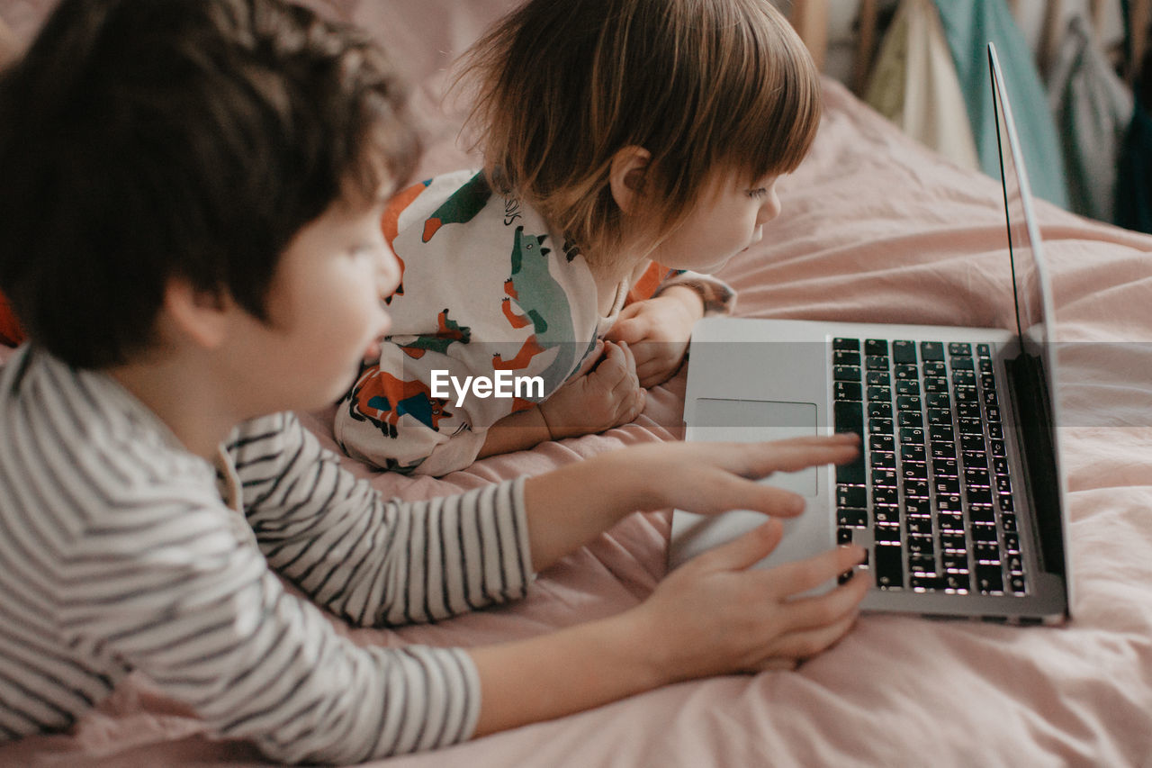 Little brother and sister lie in the bedroom on the bed and look at the laptop. high quality photo