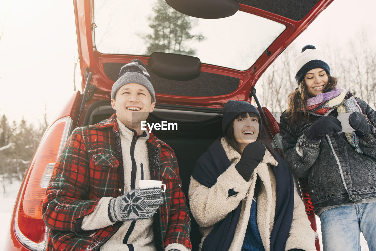 Low angle view of happy friends having coffee while resting in car trunk during winter