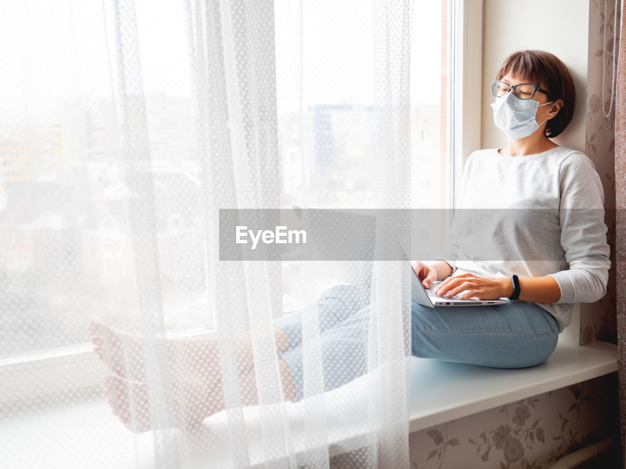 Woman in medical mask remote works from home. she sits on window sill with laptop on knees. 