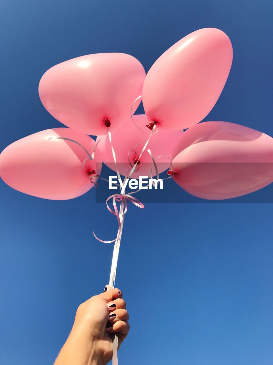 Low angle view of cropped hand holding balloons against clear blue sky