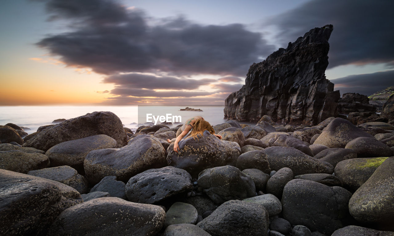 Woman lying on rocks at beach against sky during sunset