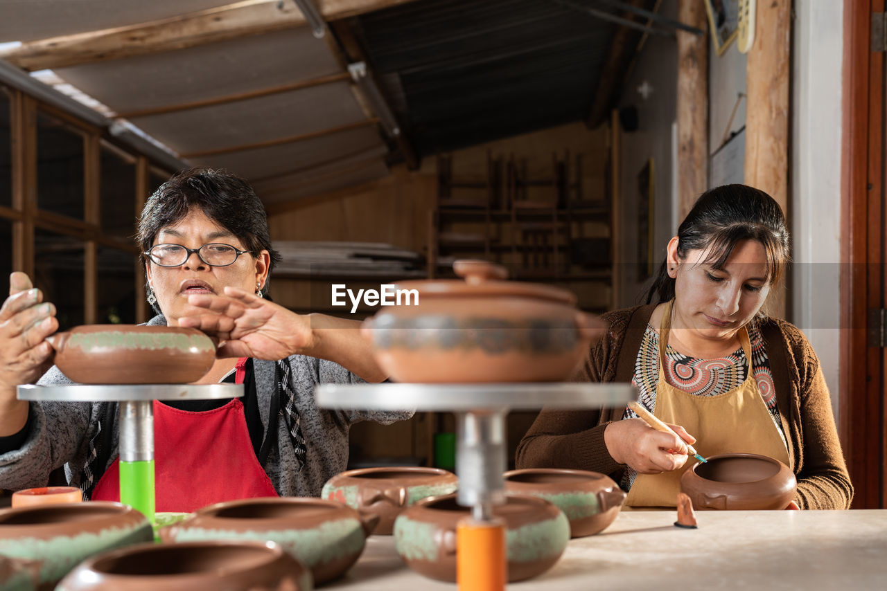 Focused peruvian female masters in aprons preparing to paint on clay pots while sitting at table in light professional workshop