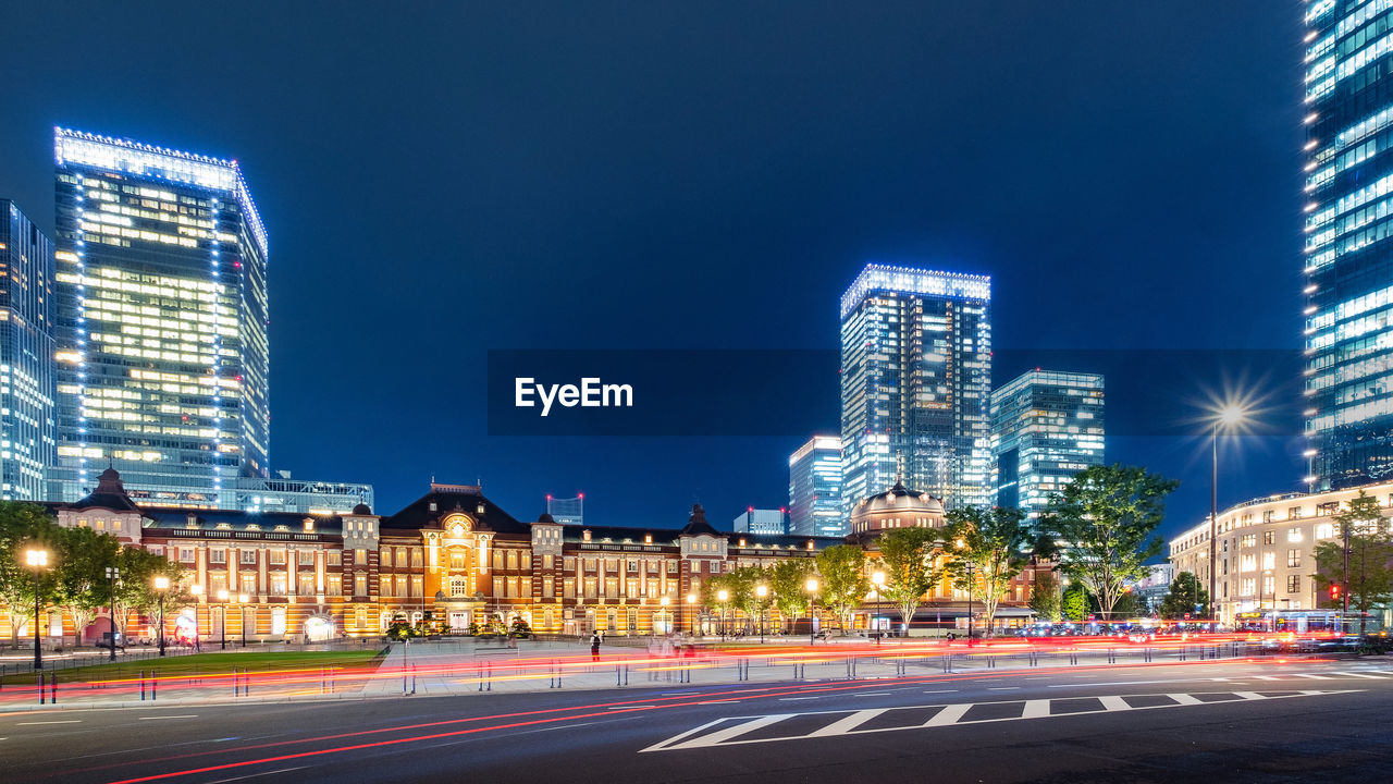 Light trails on city street by buildings against sky at night