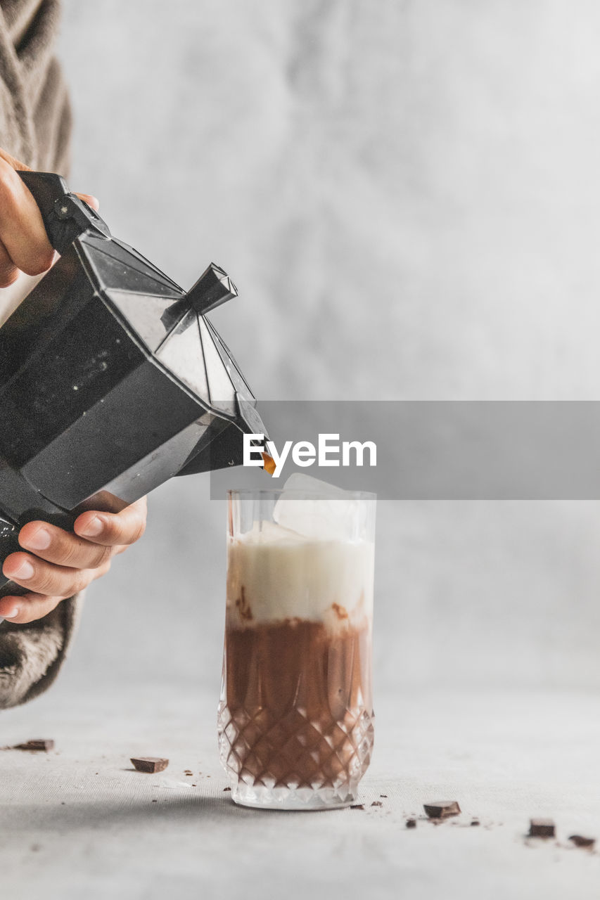 Female pouring coffee with moka pot coffee maker into dinking glass with iced mocha on table